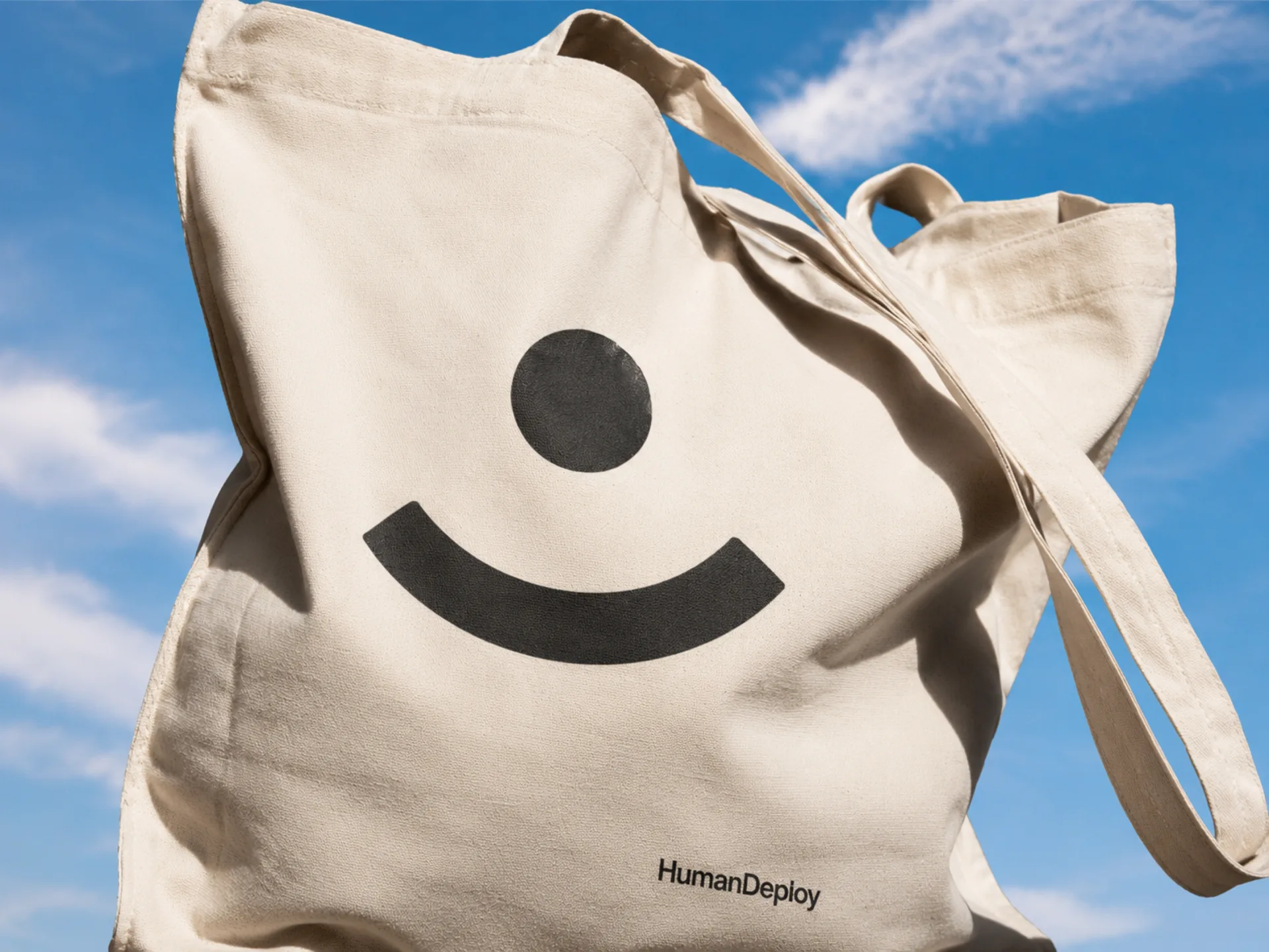 HumanDeploy — A cream cotton HumanDeploy tote against a blue sky, printed with the black smile mark and wordmark