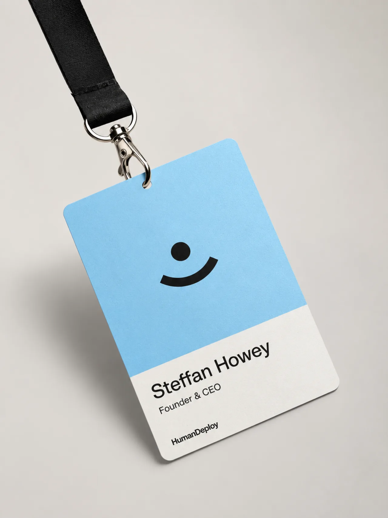 HumanDeploy — A HumanDeploy credential on a black lanyard — a sky-blue card with the smile mark above a founder name block and wordmark