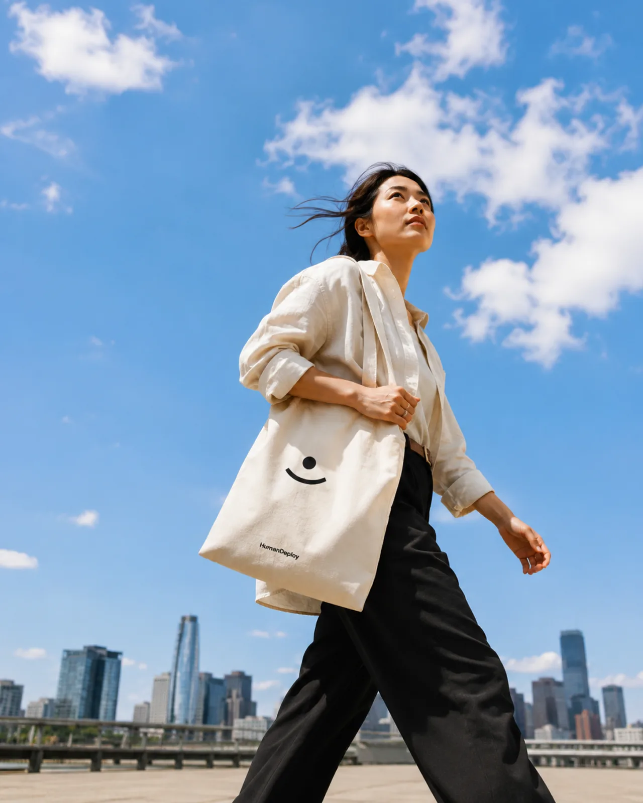 HumanDeploy — A low-angle shot of a woman crossing a plaza with a HumanDeploy tote, city towers and open sky behind her