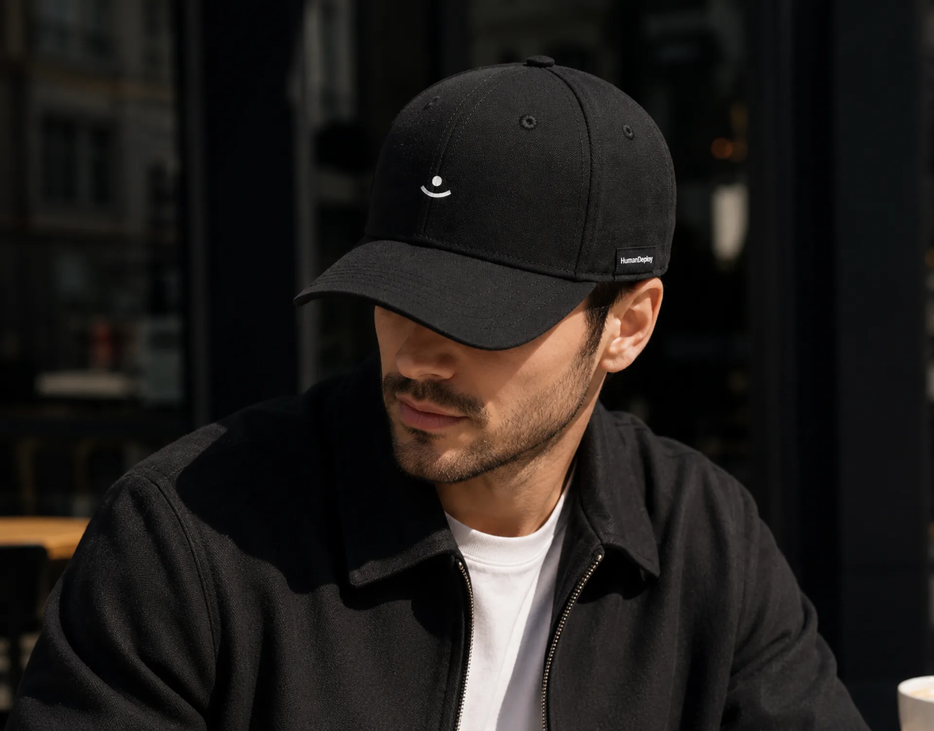 HumanDeploy — A man in a black HumanDeploy cap with a white embroidered smile mark and a woven wordmark tab, lit against a city window