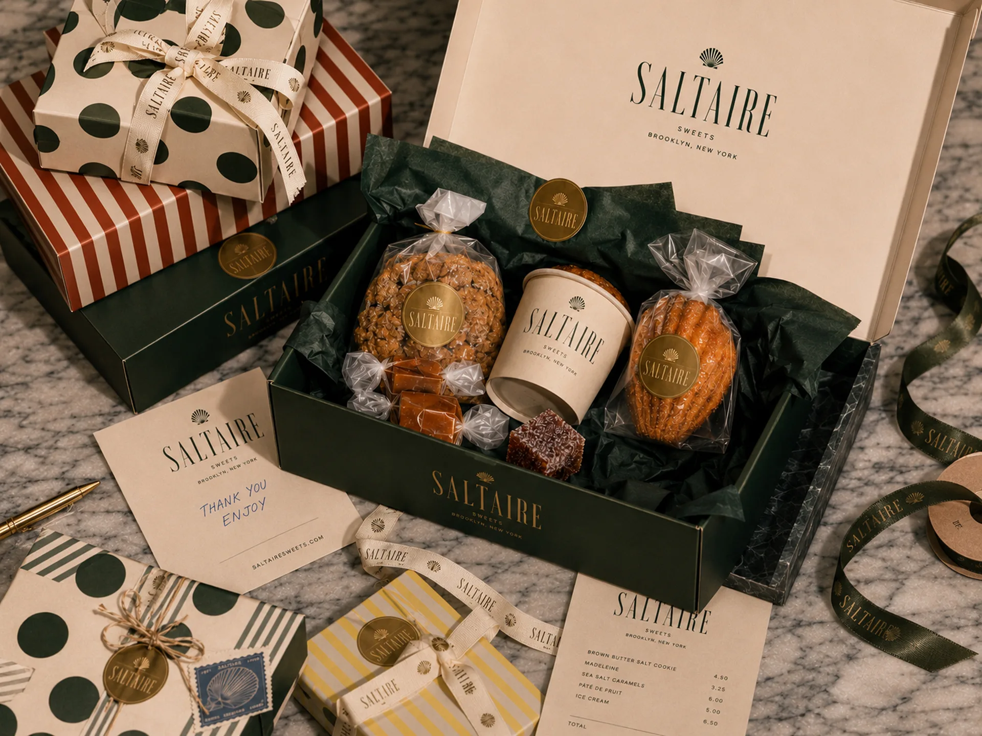 SALTAIRE — An open SALTAIRE gift box with wrapped sweets, cream cup, madeleine, cookie, printed cards, polka-dot gift wrap, and ribbons