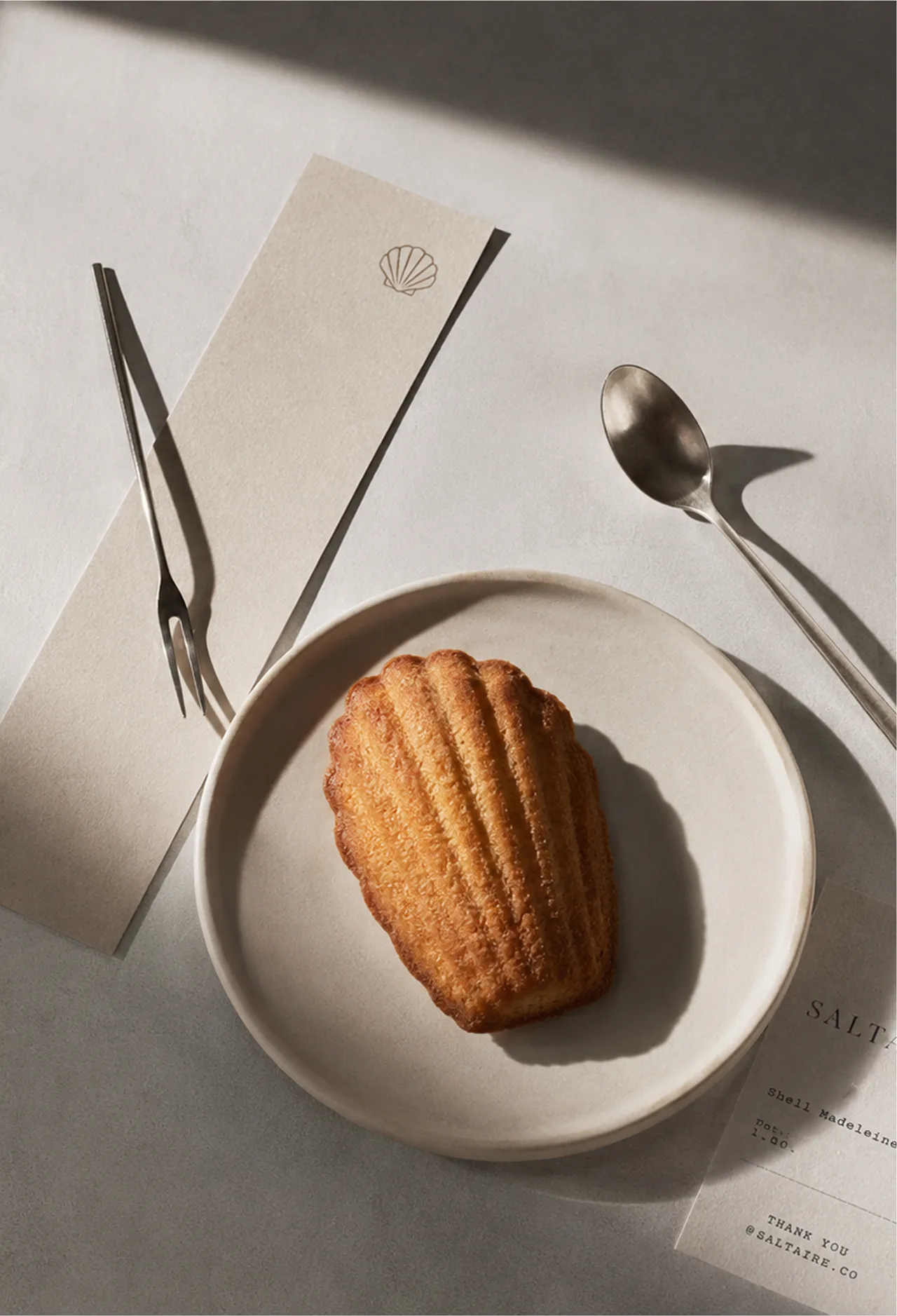 SALTAIRE — A single golden madeleine on a ceramic plate, surrounded by a receipt slip, spoon, fork, hard shadows, and quiet shop ephemera