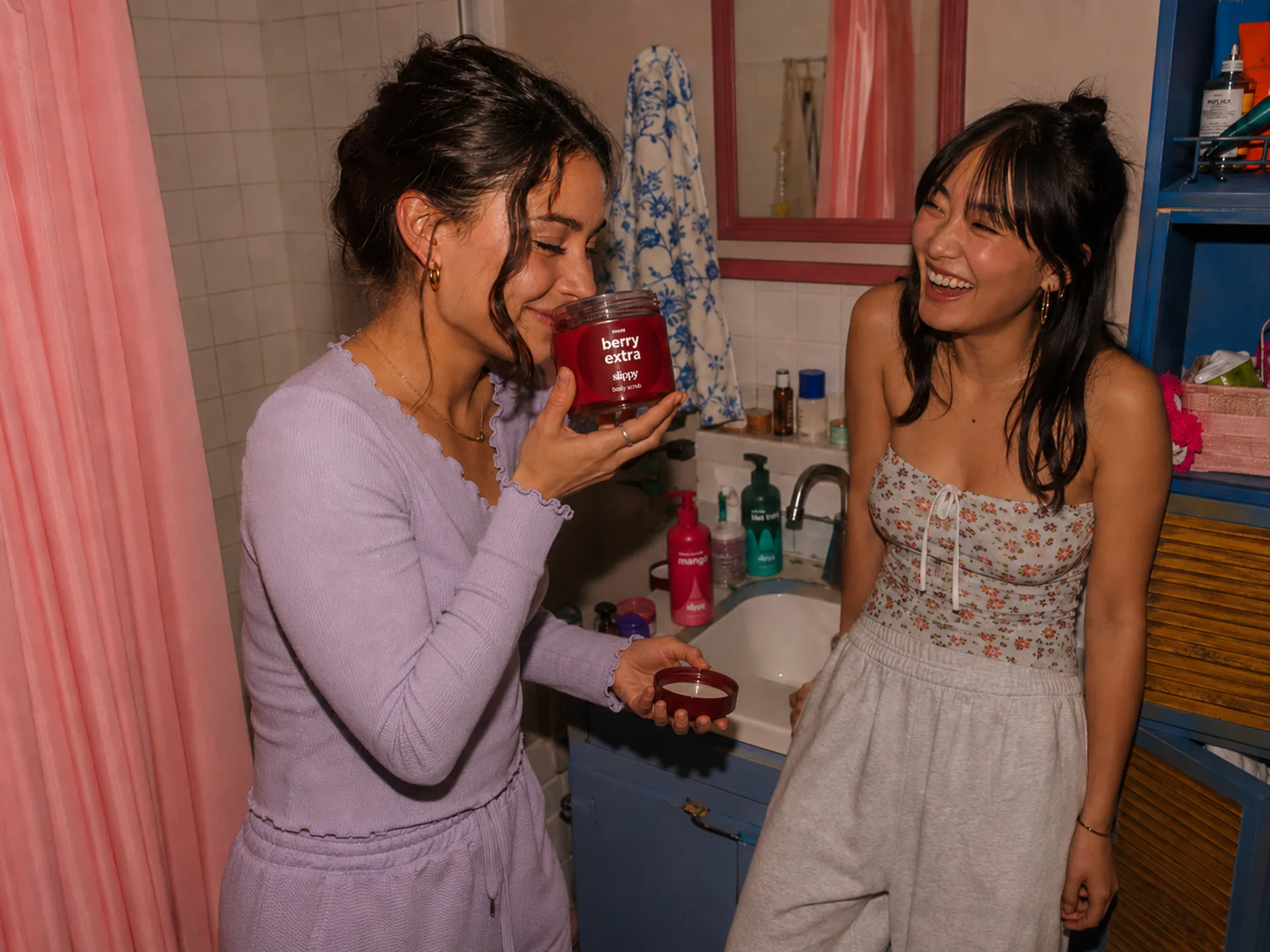 Slippy — Friends in a small bathroom passing around berry extra body scrub with a candid getting-ready energy