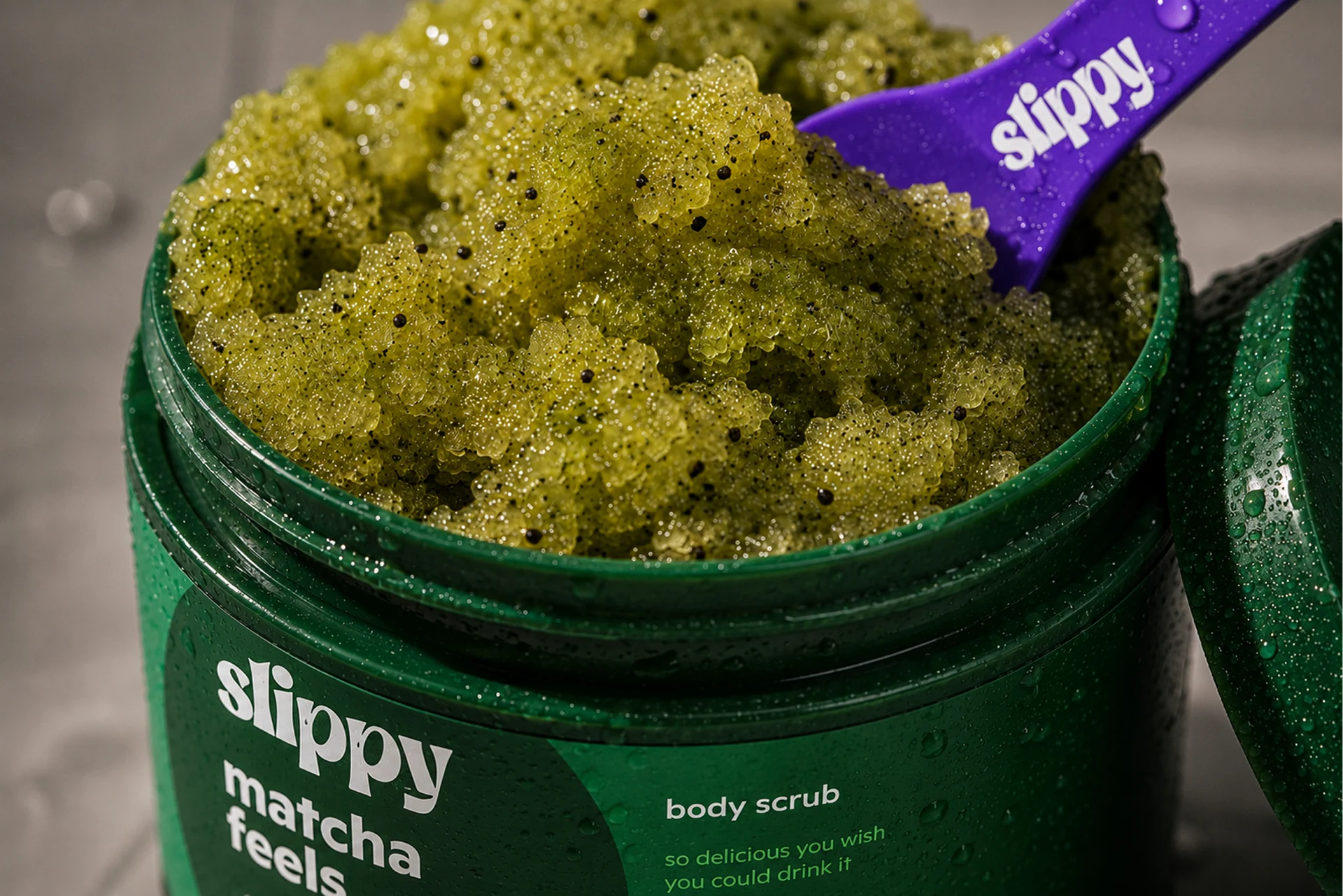 Slippy — Close detail of matcha feels body scrub with granular green texture, water beads, and a branded scoop