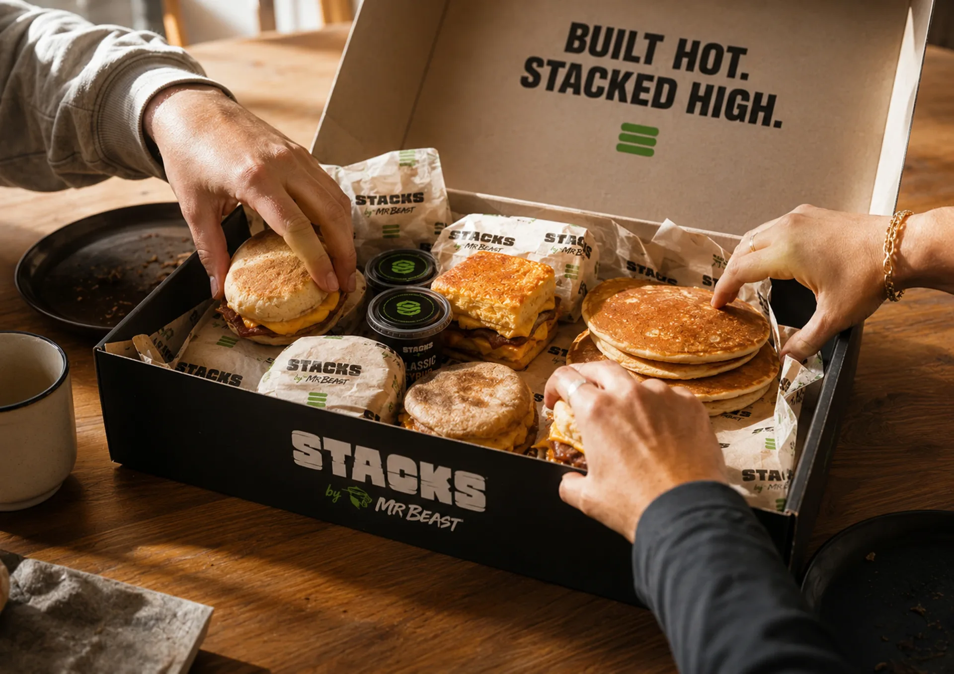 Stacks — Hands sharing a Stacks breakfast kit with pancake stack, sandwich, sauces, coffee, and branded wrap