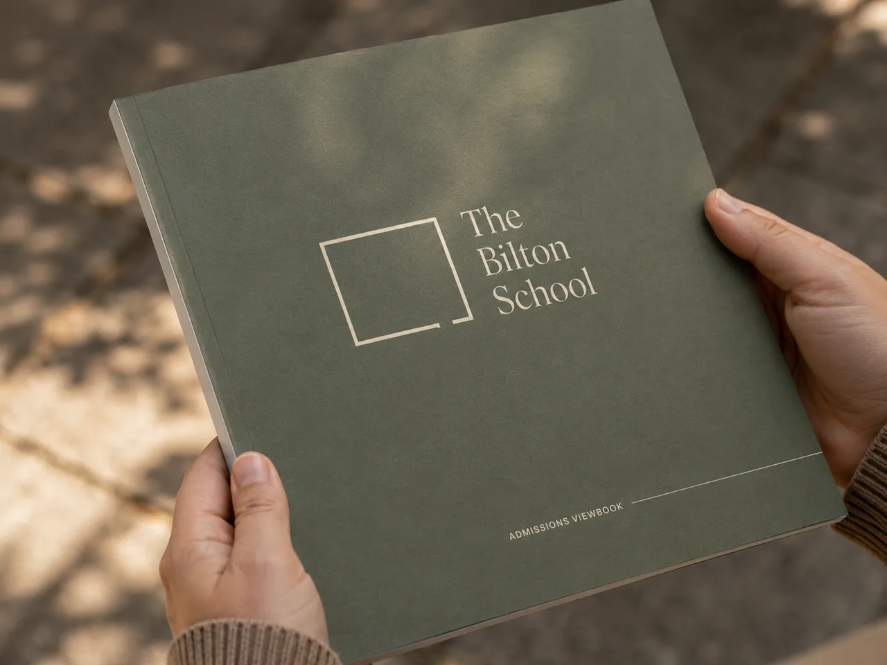 The Bilton School — Admissions viewbook held in hand on a sunlit campus walk with olive cover and serif wordmark